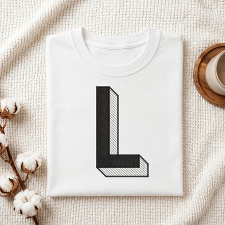 A folded white T-shirt with a large black block letter 'L' printed on the front, styled on a textured cream fabric with cotton stems and a cup in the background.