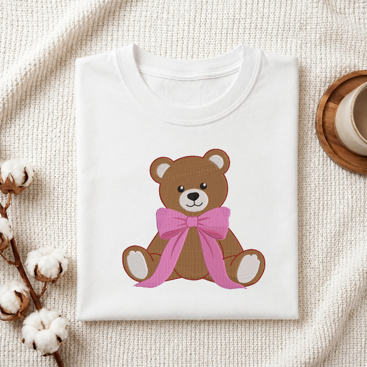 A folded white T-shirt laid flat on a textured cream fabric background, featuring a brown teddy bear graphic with a pink bow on the front. Cotton stems and part of a wooden cup are visible as styled props.