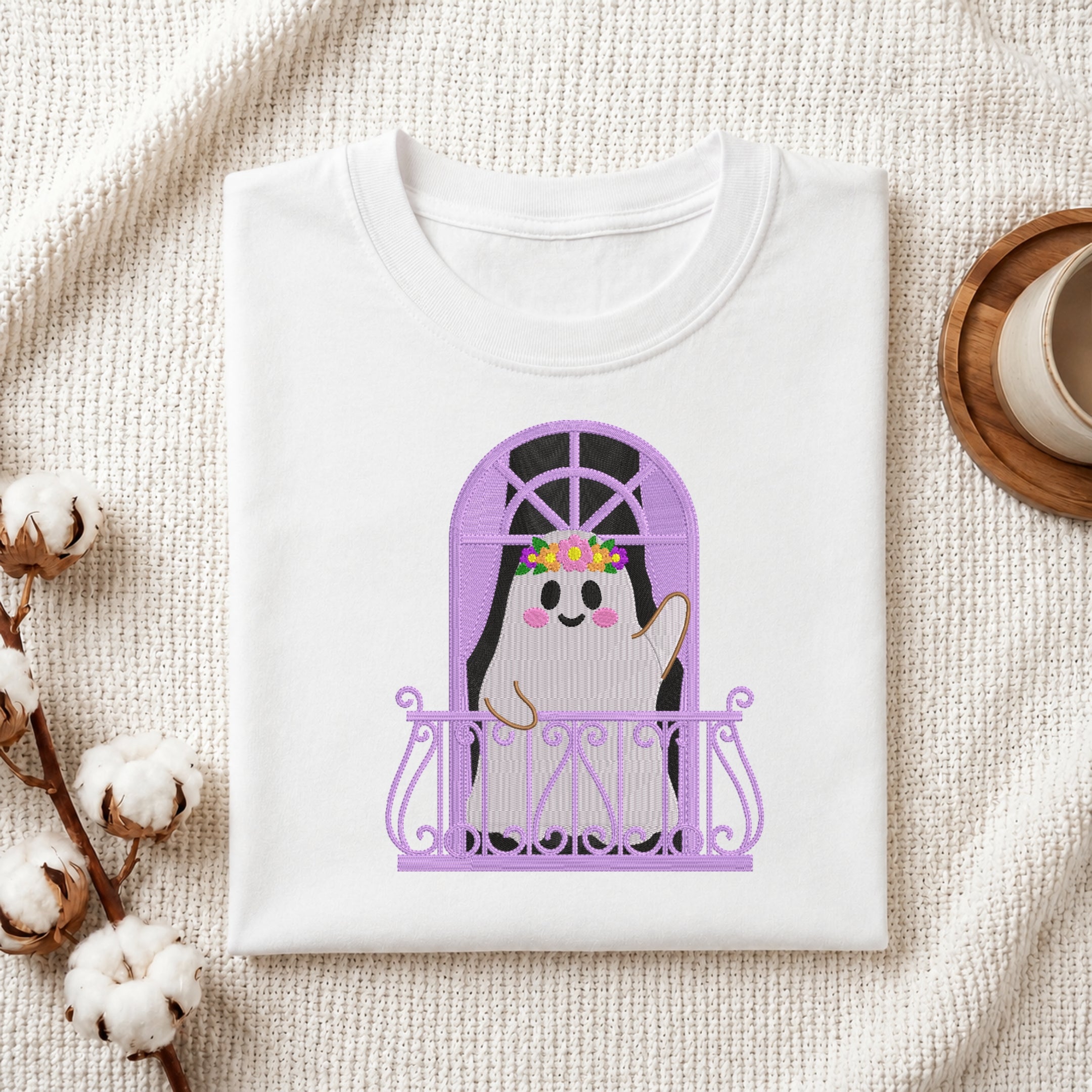 A folded white crewneck t-shirt displayed flat on a textured cream fabric background, decorated with cotton bolls on the left and a wooden cup on the right. The t-shirt features a graphic of a cute cartoon ghost peeking out from behind a purple arched window with a lavender railing.