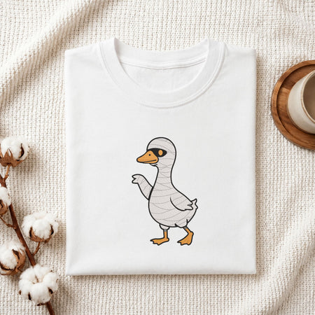 Folded white cotton t-shirt mockup featuring a cute duck in a mummy costume Halloween machine embroidery design, styled with cotton stems and a wooden cup