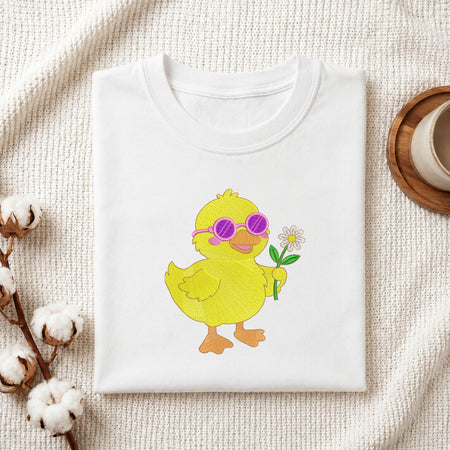 A folded white T-shirt laid flat on a textured cream fabric background, featuring a cartoon yellow duckling wearing pink sunglasses and holding a small white flower. Cotton bolls and part of a wooden cup are styled around the shirt.