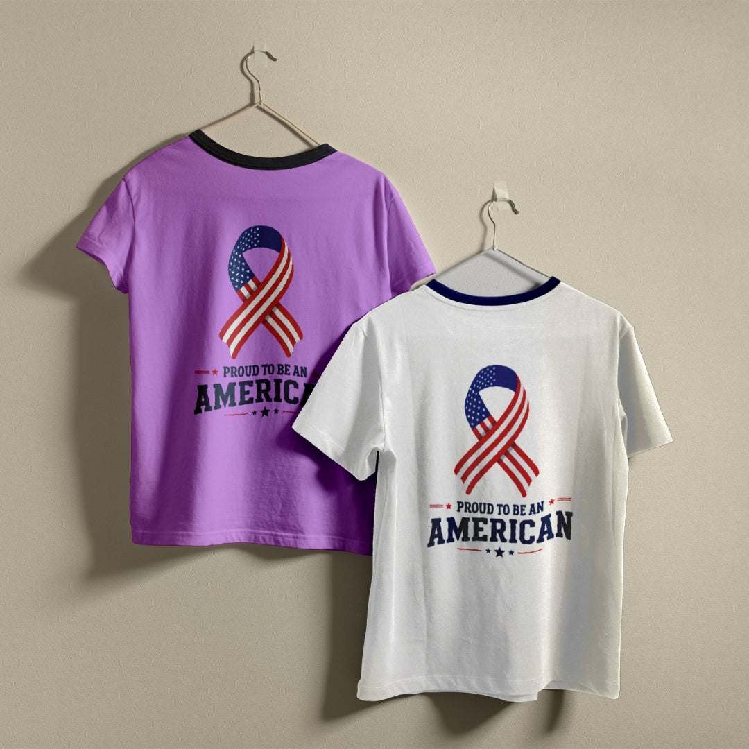 Two t-shirts hanging on hangers, one purple and one white, both printed with an American flag ribbon graphic and the text "Proud to be an American."