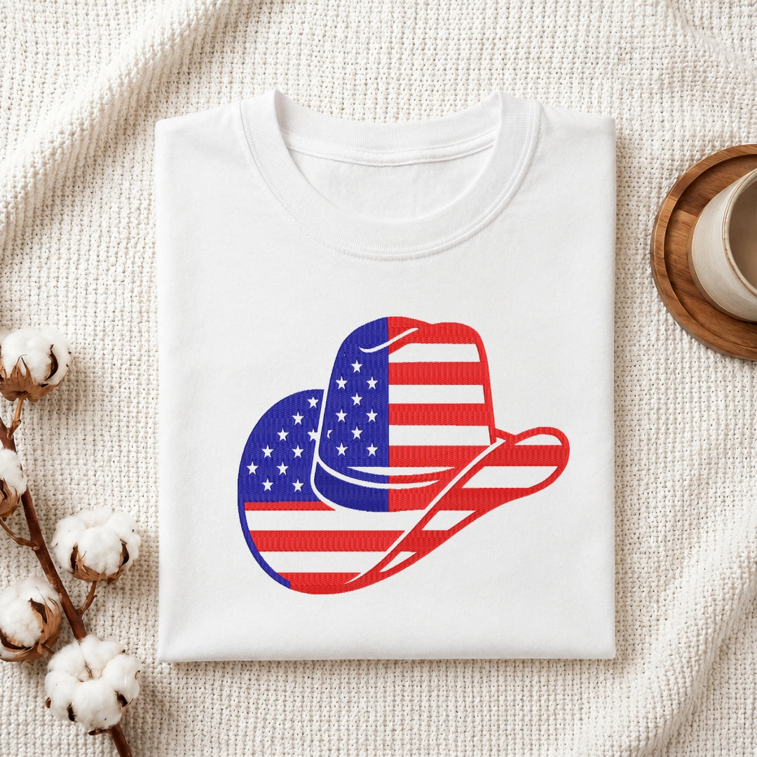 A folded white crewneck t-shirt with a graphic of a cowboy hat filled with the American flag pattern, styled on a textured cream fabric with cotton bolls and a wooden cup.