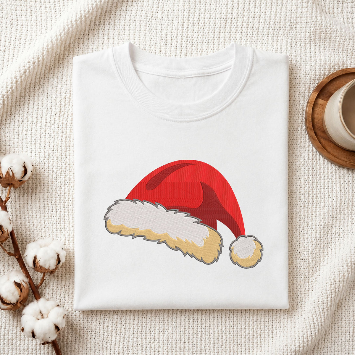 Folded white cotton t-shirt mockup featuring a Santa hat embroidery design, styled with cotton stems and a wooden cup.