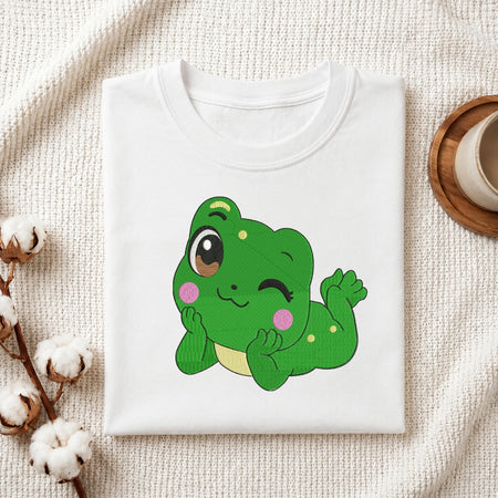 A folded white crewneck t-shirt featuring a cute cartoon green frog graphic, styled on a textured cream fabric background with cotton bolls and a wooden cup.