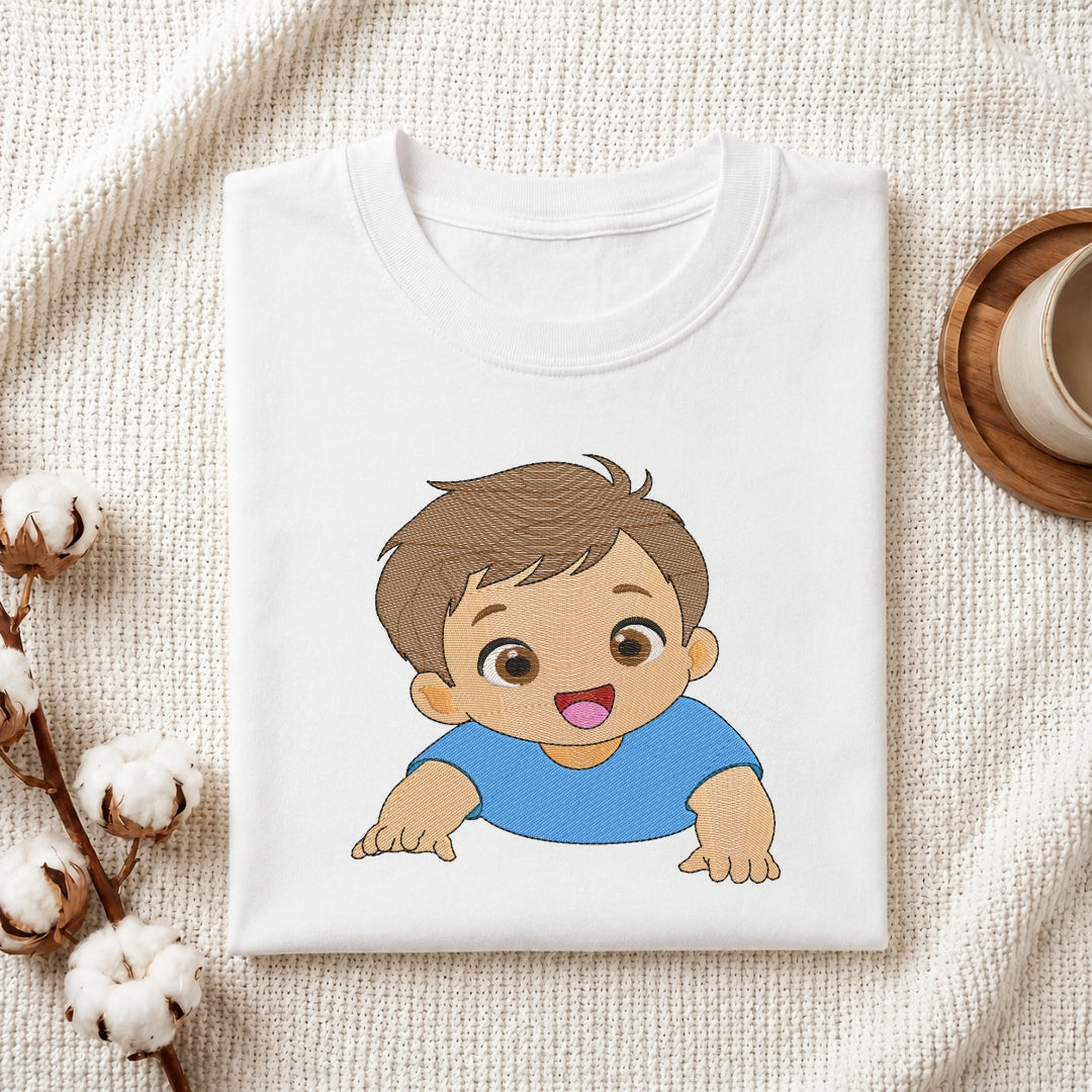 A folded white crewneck t-shirt featuring a cartoon illustration of a smiling baby boy with brown hair and a blue shirt, displayed on a textured cream fabric background with decorative cotton stems and a wooden cup.