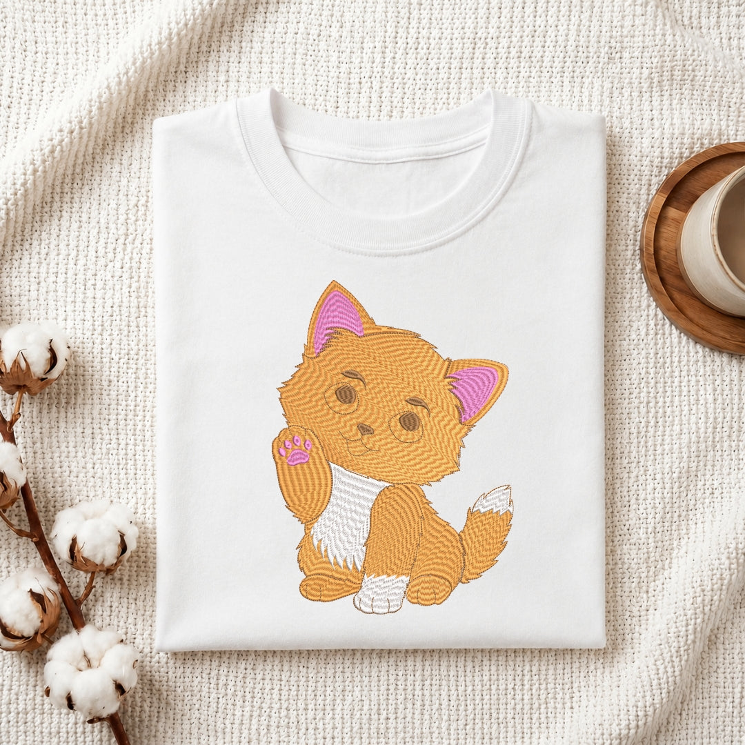 A white folded t-shirt featuring a detailed, embroidered orange kitten with pink ears, placed on a textured white fabric with cotton sprigs and a wooden coaster.