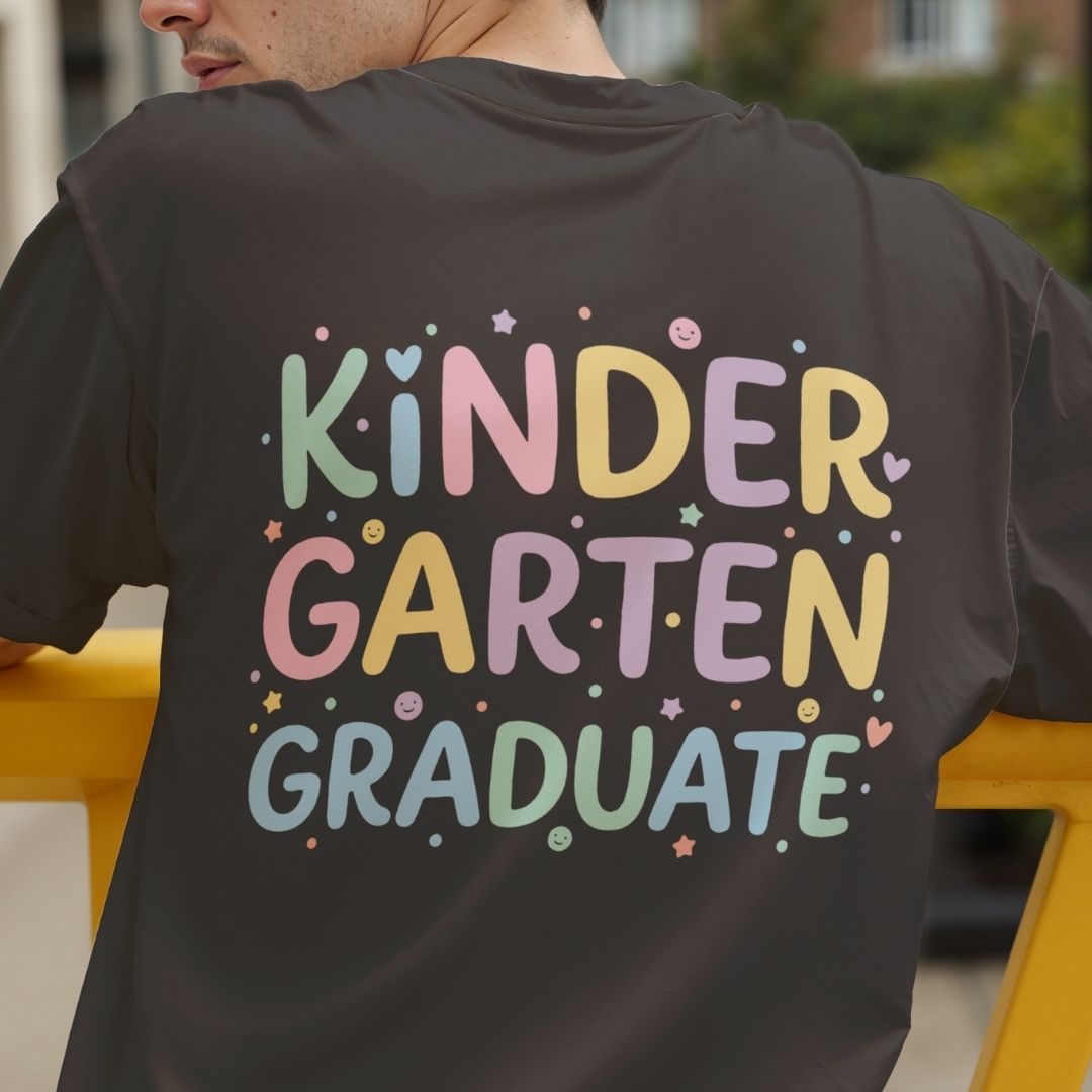 A person wearing a dark charcoal gray t-shirt, viewed from behind, featuring a colorful pastel graphic that reads 'KINDER GARTEN GRADUATE' surrounded by small stars and dots.