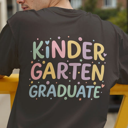 A person wearing a dark charcoal gray t-shirt, viewed from behind, featuring a colorful pastel graphic that reads 'KINDER GARTEN GRADUATE' surrounded by small stars and dots.