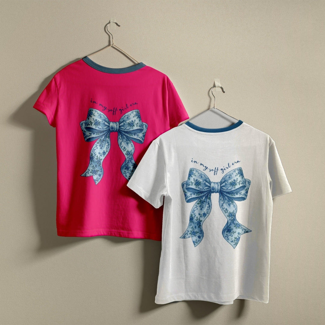 Two t-shirts hanging on hangers, one hot pink and one white, both printed with a blue floral bow graphic and the text "in my soft girl era."