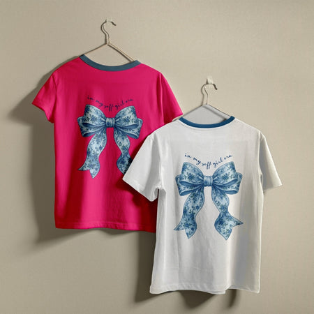 Two t-shirts hanging on hangers, one hot pink and one white, both printed with a blue floral bow graphic and the text "in my soft girl era."