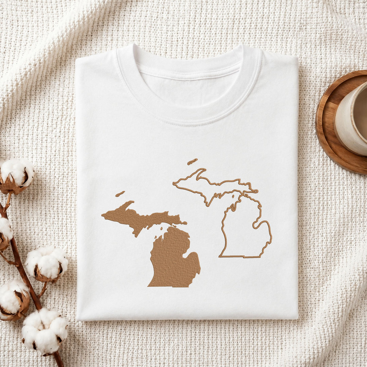 A folded white T-shirt with a brown outline graphic of the state of Michigan on the front, styled on a textured cream fabric with cotton bolls and a wooden cup in the background.