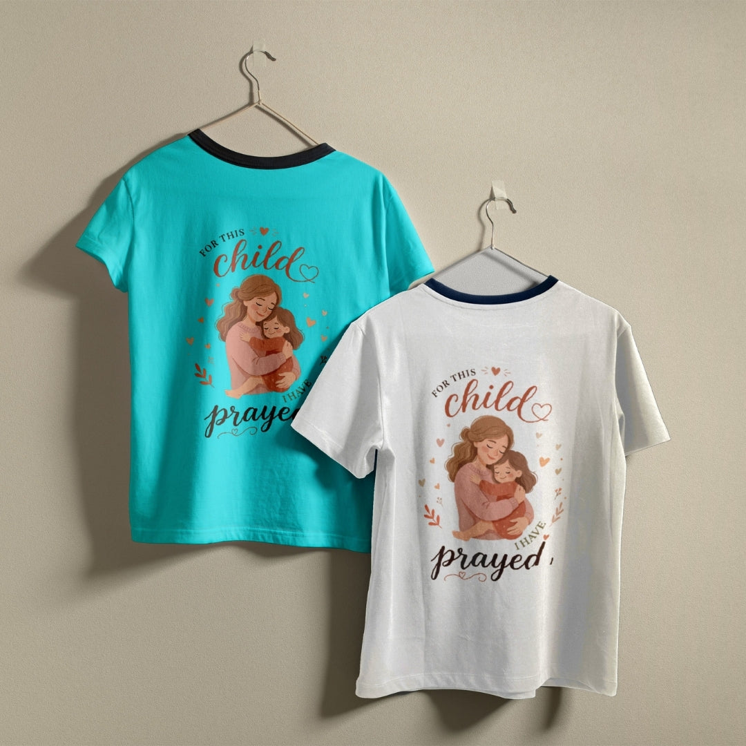 Two t-shirts hanging on hangers, one purple and one white, both printed with a mother-and-child illustration and the text "For this child I have prayed."