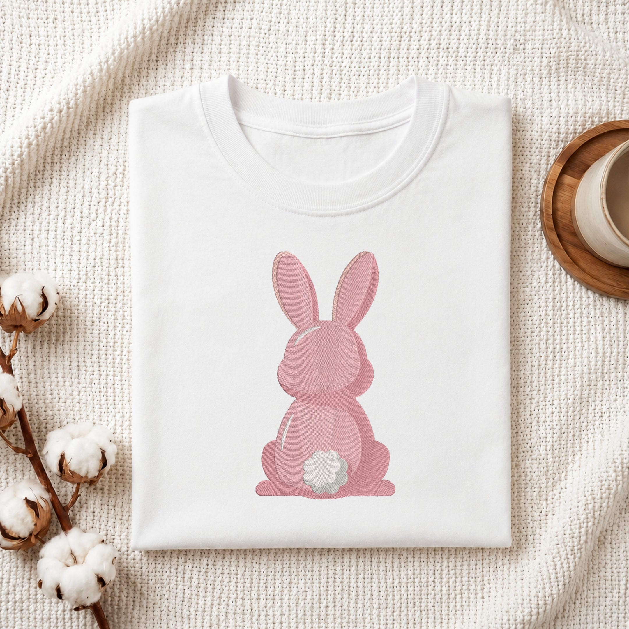 A folded white T-shirt with a pink bunny graphic sitting on a textured cream fabric background, styled with cotton bolls and a wooden cup on a coaster.