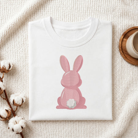 A folded white T-shirt with a pink bunny graphic sitting on a textured cream fabric background, styled with cotton bolls and a wooden cup on a coaster.