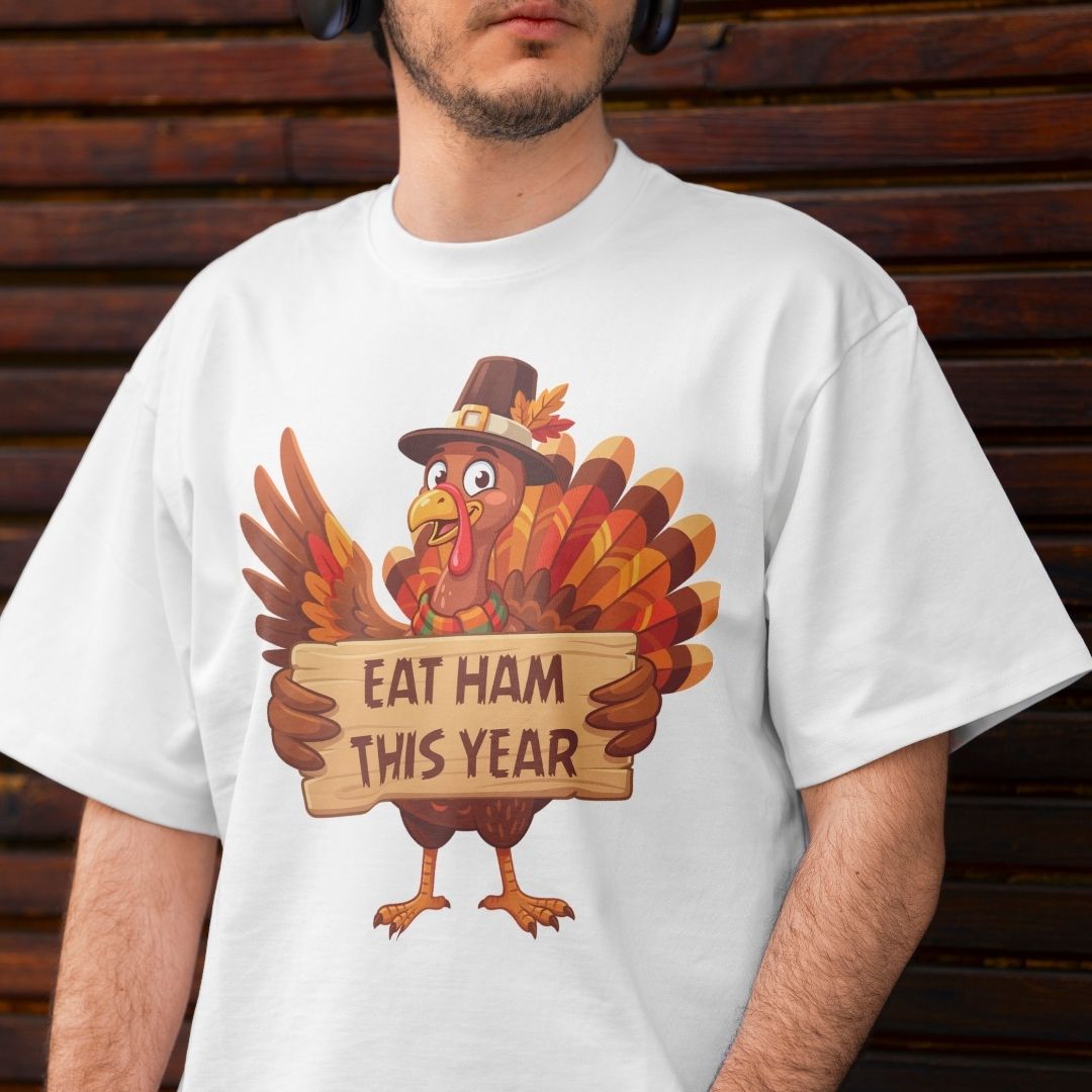 A man wearing a white t-shirt with a cartoon turkey graphic holding a sign that reads 'EAT HAM THIS YEAR', set against a dark wooden background.