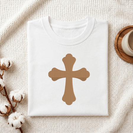 A folded white t-shirt with a brown cross graphic printed on the chest, styled on a textured cream blanket with cotton bolls and a wooden cup in the background.