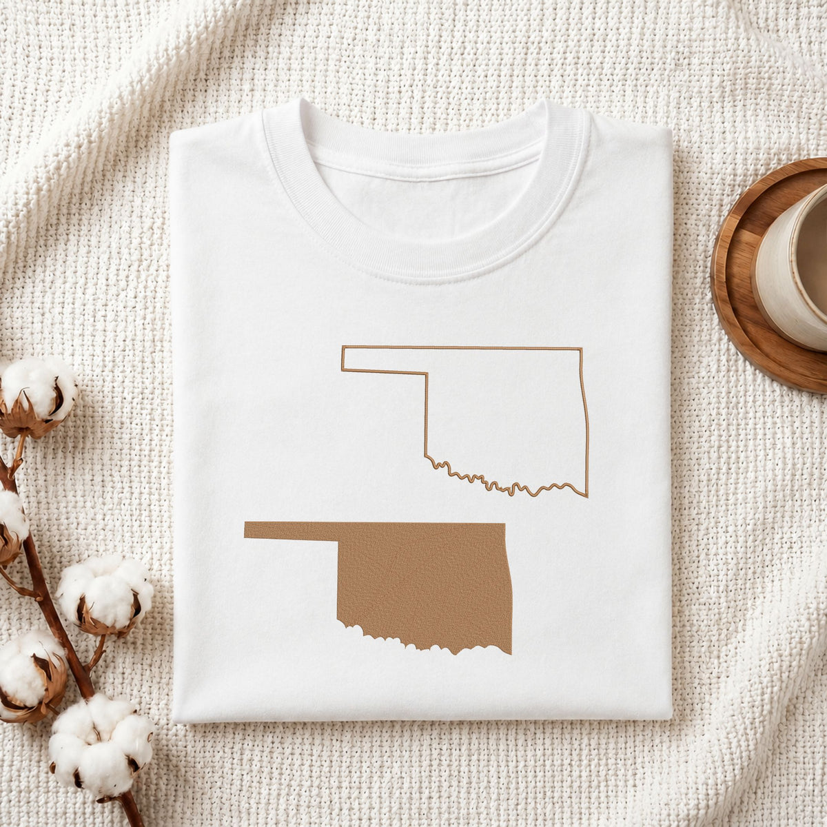 A folded white T-shirt laid flat on a textured cream fabric background, featuring a brown outline and filled silhouette of Oklahoma state. Cotton bolls and part of a wooden cup are visible as styled props.