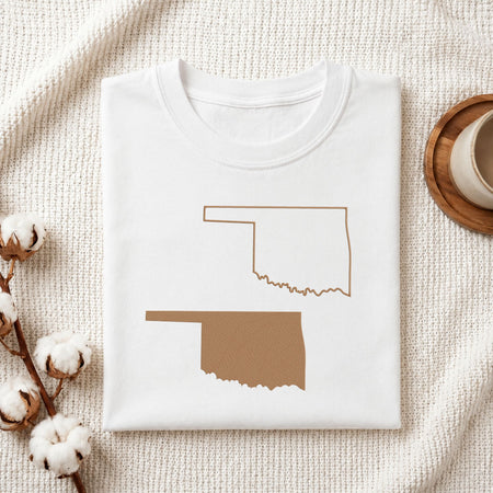 A folded white T-shirt laid flat on a textured cream fabric background, featuring a brown outline and filled silhouette of Oklahoma state. Cotton bolls and part of a wooden cup are visible as styled props.