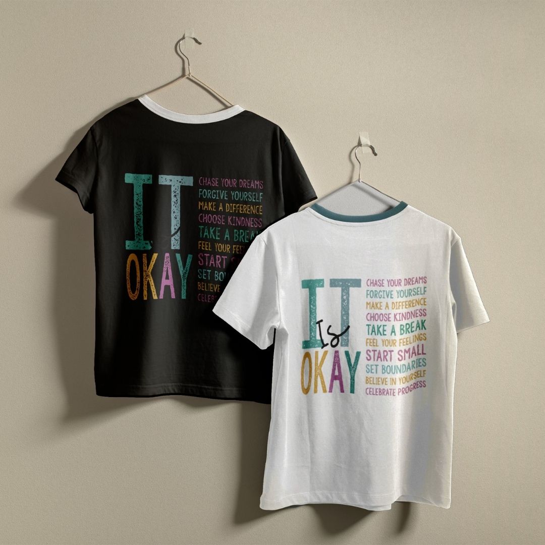 Two graphic t-shirts hanging on hangers against a plain beige wall. The left shirt is black with a white collar and features a colorful 'IT is OKAY' text design.