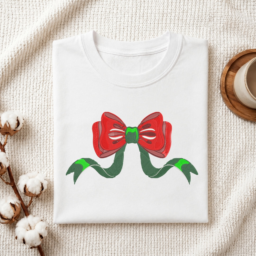 A folded white crewneck t-shirt featuring a graphic of a red bow with green ribbons, styled on a textured cream fabric background with cotton bolls and a coffee cup.