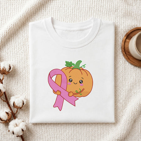 Folded white cotton t-shirt mockup featuring a cute pumpkin with a pink awareness ribbon machine embroidery design, styled with cotton stems and a wooden cup.