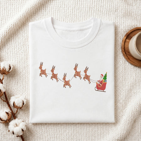 A folded white crewneck t-shirt with a graphic of Santa Claus in a sleigh pulled by four brown reindeer, styled on a textured cream knit blanket with cotton bolls and a coffee cup.