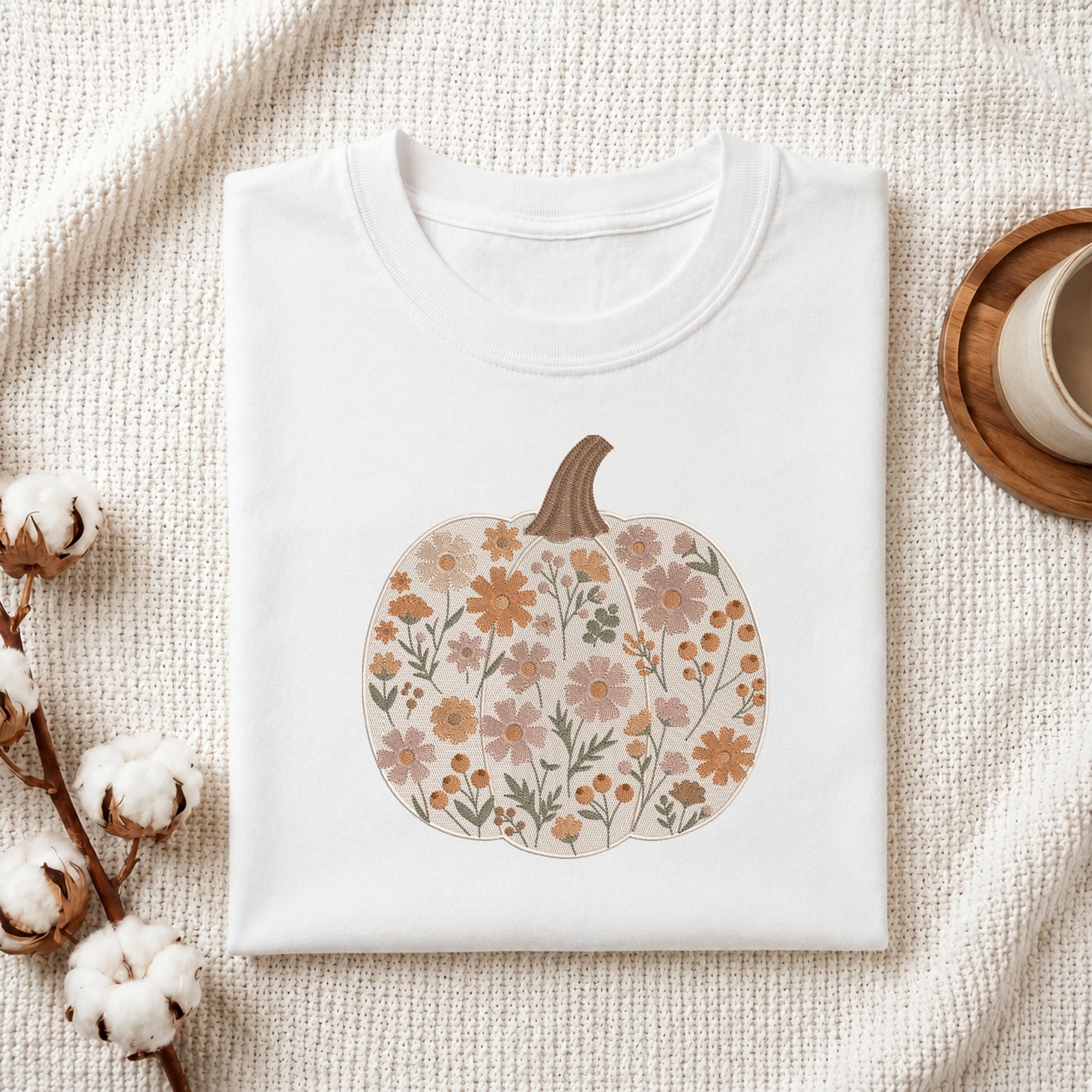 A folded white crewneck t-shirt with a floral pumpkin graphic, styled on a textured cream fabric with cotton bolls and a wooden cup on a coaster.