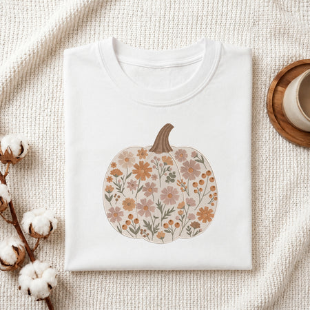 A folded white crewneck t-shirt with a floral pumpkin graphic, styled on a textured cream fabric with cotton bolls and a wooden cup on a coaster.