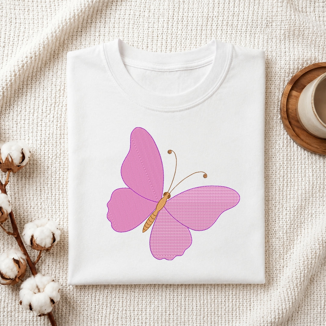 A folded white crewneck t-shirt featuring a pink butterfly graphic on the front, styled on a textured cream fabric background with cotton bolls and a wooden cup.