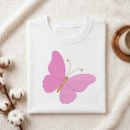 A folded white crewneck t-shirt featuring a pink butterfly graphic on the front, styled on a textured cream fabric background with cotton bolls and a wooden cup.