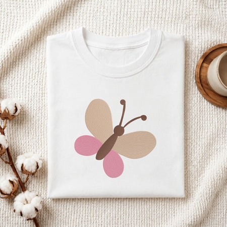 Folded white cotton t-shirt mockup featuring a minimalist butterfly machine embroidery design, styled with cotton stems and a wooden cup
