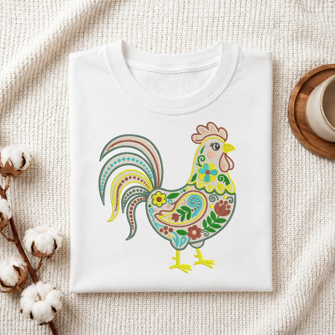 A folded white crewneck t-shirt featuring a colorful, ornate rooster graphic on the front, styled on a textured cream fabric with cotton bolls and a wooden cup in the background.