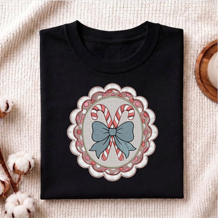 A folded black crewneck t-shirt with a festive Christmas graphic, displayed on a textured beige fabric background with decorative cotton bolls and a wooden tray.