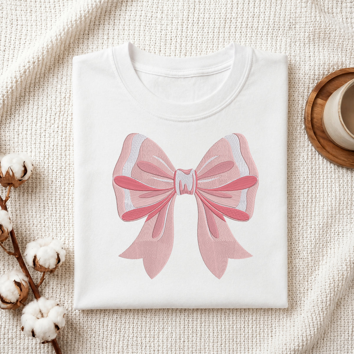 A folded white T-shirt with a large pink bow graphic on the front, styled on a textured cream fabric with cotton bolls and a small cup in the background.