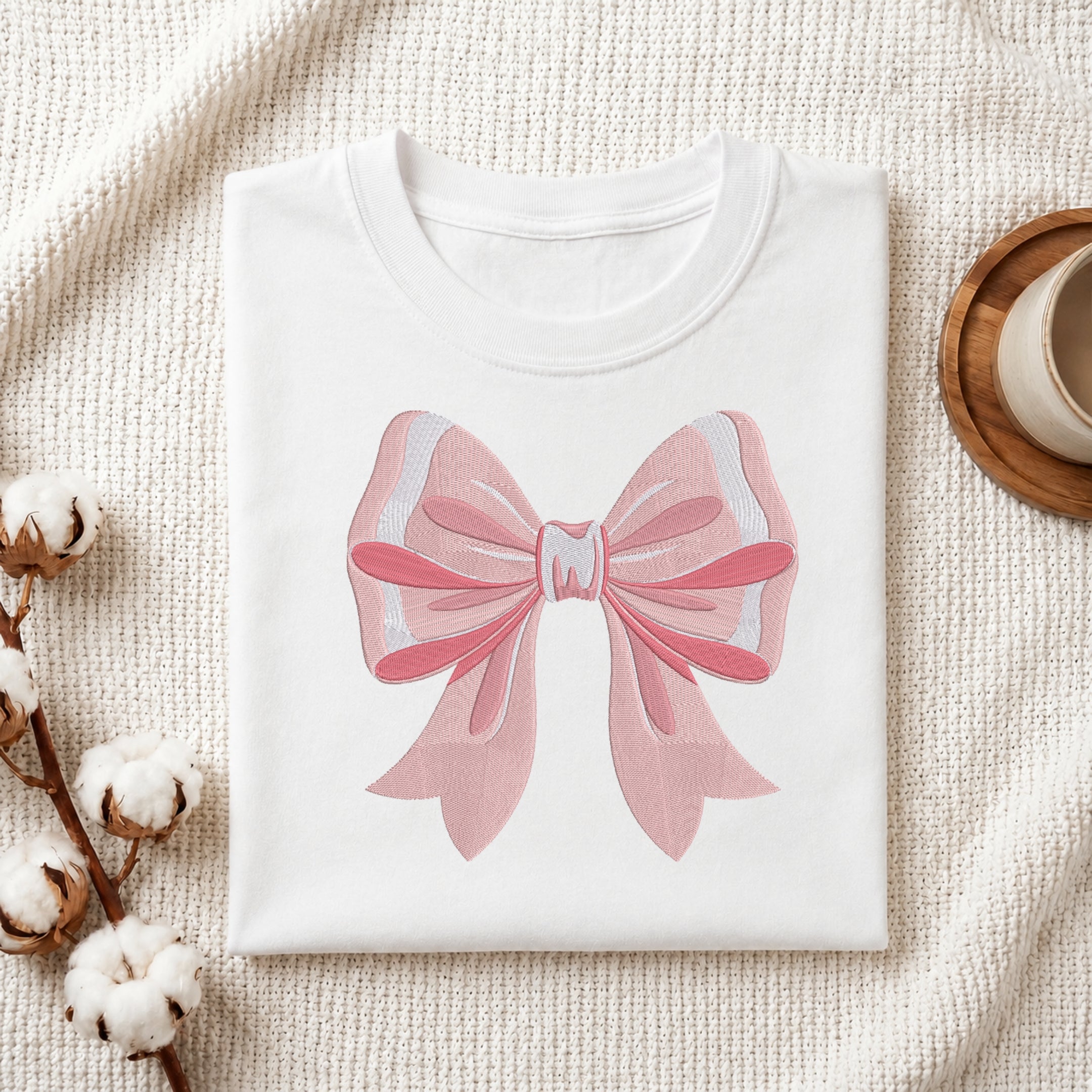 A folded white T-shirt with a large pink bow graphic on the front, styled on a textured cream fabric with cotton bolls and a small cup in the background.
