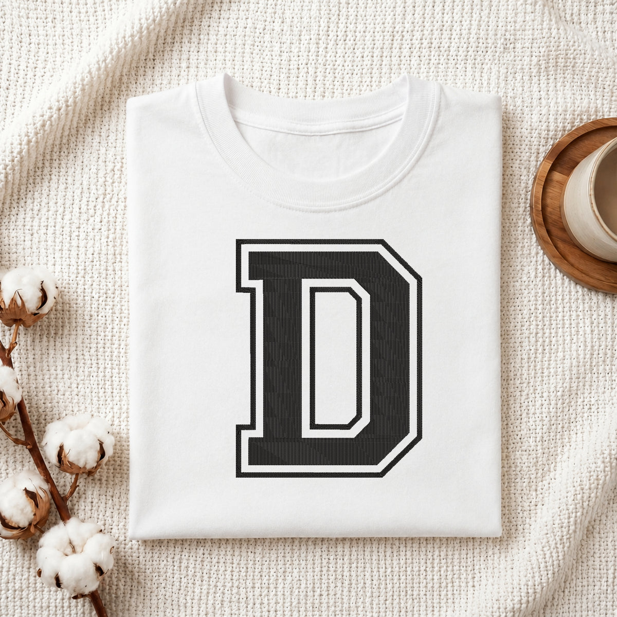 A folded white t-shirt with a large black and white block letter 'D' printed on the front, styled on a textured cream knit blanket with cotton bolls and a cup in the background.