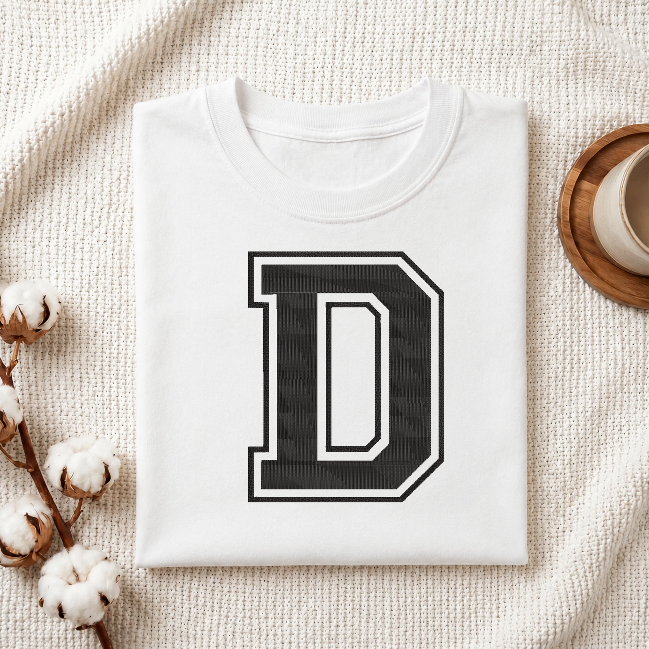 A folded white t-shirt with a large black and white block letter 'D' printed on the front, styled on a textured cream knit blanket with cotton bolls and a cup in the background.