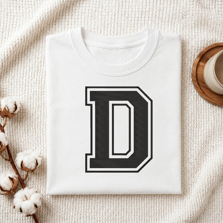 A folded white t-shirt with a large black and white block letter 'D' printed on the front, styled on a textured cream knit blanket with cotton bolls and a cup in the background.