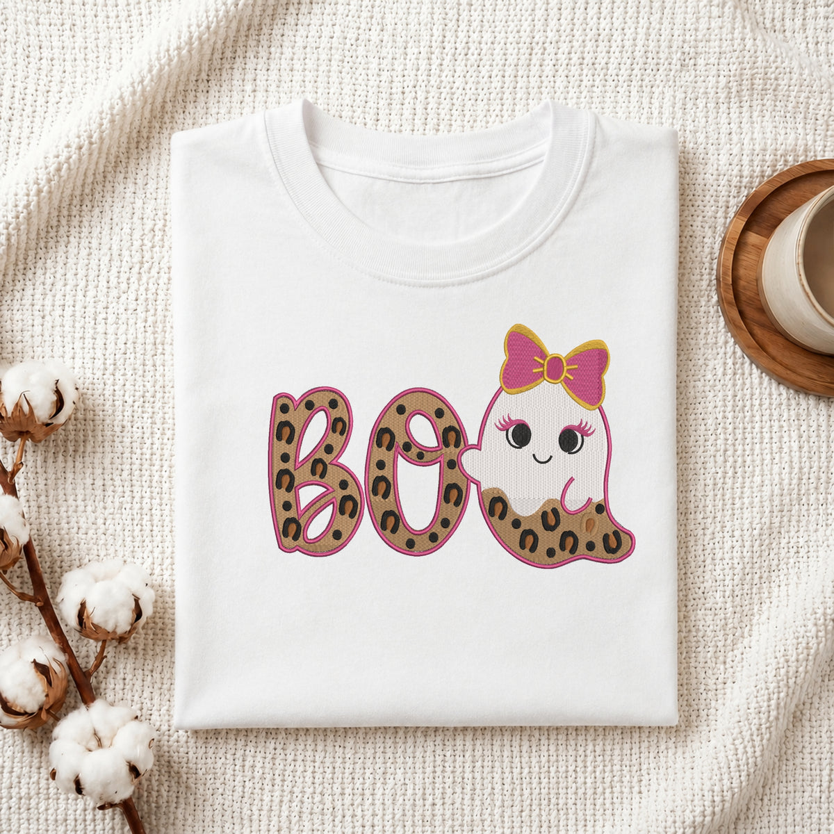 A neatly folded white t-shirt featuring the embroidered leopard print "BOO" and cute ghost design, displayed on a textured white knit background with cotton stems and a wooden cup.