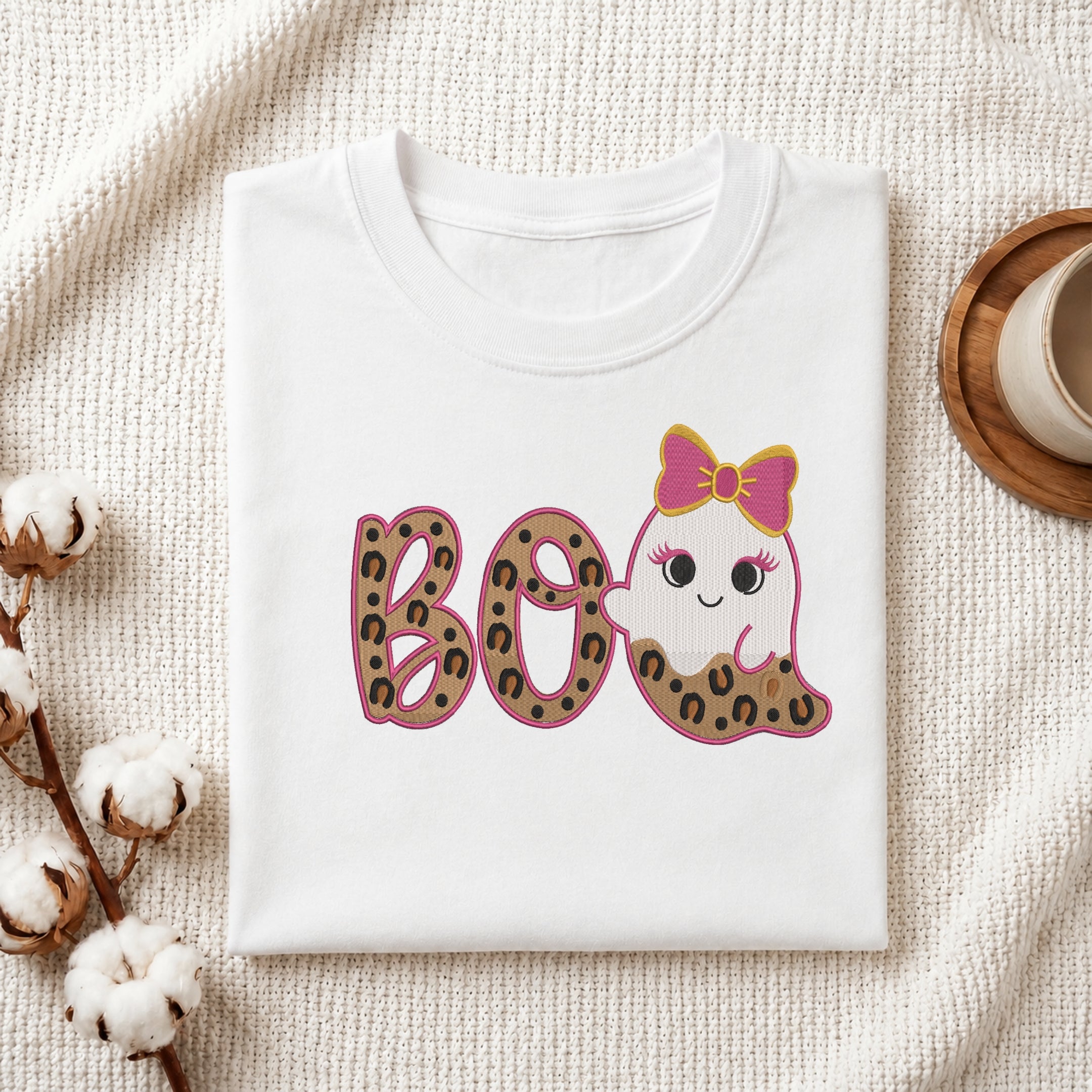A neatly folded white t-shirt featuring the embroidered leopard print "BOO" and cute ghost design, displayed on a textured white knit background with cotton stems and a wooden cup.