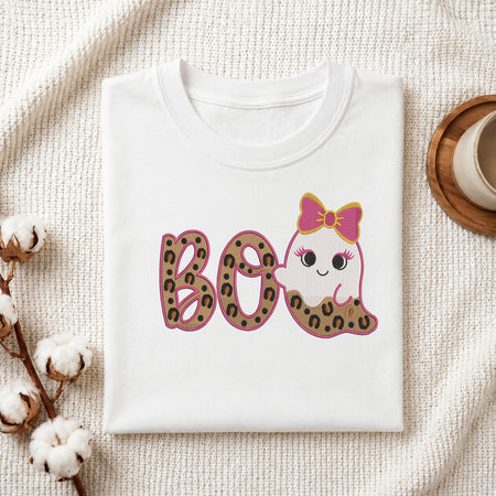 A neatly folded white t-shirt featuring the embroidered leopard print "BOO" and cute ghost design, displayed on a textured white knit background with cotton stems and a wooden cup.