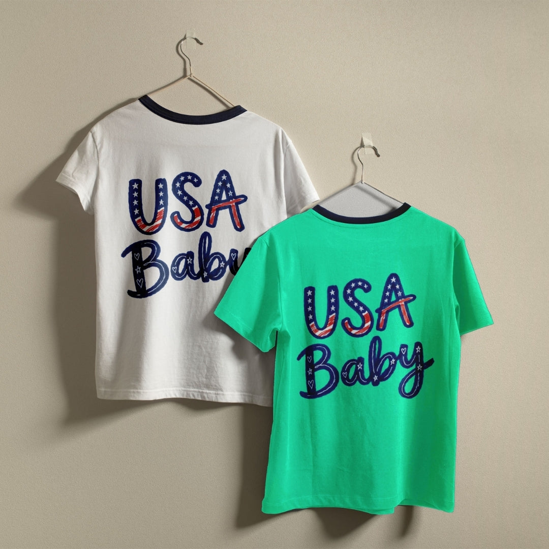 Two ringer t-shirts hanging on hangers against a plain beige wall. The left shirt is white with a navy collar and the back features 'USA Baby' in an American flag pattern.