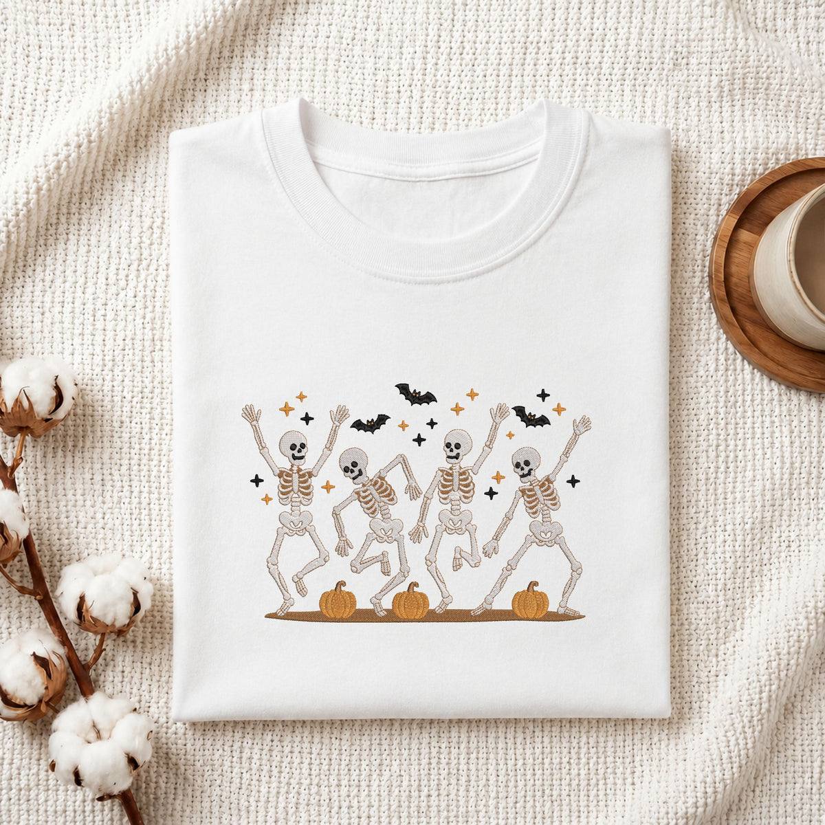Folded white cotton t-shirt mockup featuring a cute dancing skeletons Halloween machine embroidery design, styled with cotton stems and a wooden cup