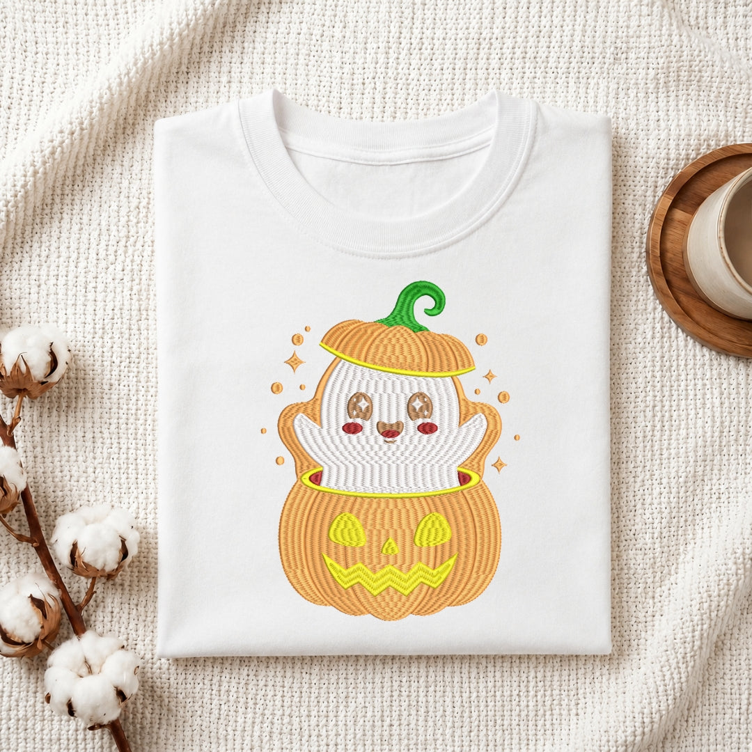 A folded white T-shirt with a cute embroidered Halloween graphic of a ghost inside a jack-o'-lantern, styled on a textured cream blanket with cotton bolls and a cup in the background.