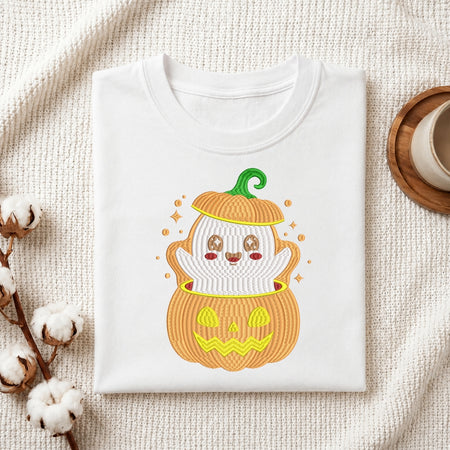 A folded white T-shirt with a cute embroidered Halloween graphic of a ghost inside a jack-o'-lantern, styled on a textured cream blanket with cotton bolls and a cup in the background.