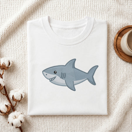A folded white crewneck t-shirt featuring a cartoon-style blue and gray shark graphic on the front, styled on a textured cream fabric with cotton bolls and a wooden cup.