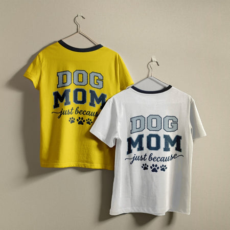 Two t-shirts hanging on a wall, one bright yellow and one white, both featuring the retro collegiate "Dog Mom just because" design with dog paw prints.
