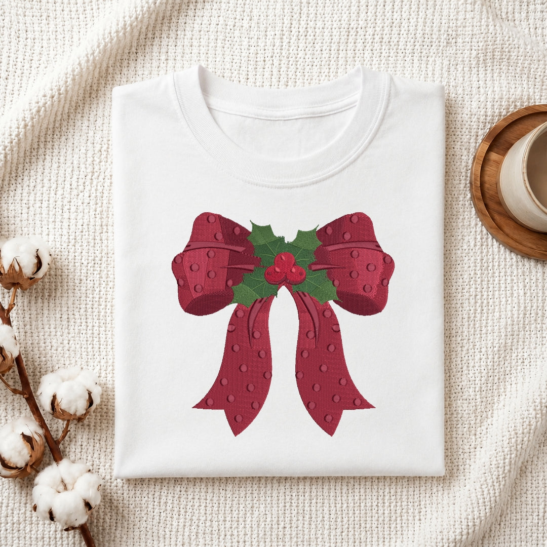 A folded white crewneck t-shirt featuring a festive graphic of a red bow with green holly leaves and red berries, styled on a textured cream fabric with cotton stems and a wooden cup in the background.