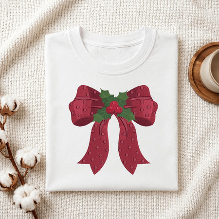 A folded white crewneck t-shirt featuring a festive graphic of a red bow with green holly leaves and red berries, styled on a textured cream fabric with cotton stems and a wooden cup in the background.